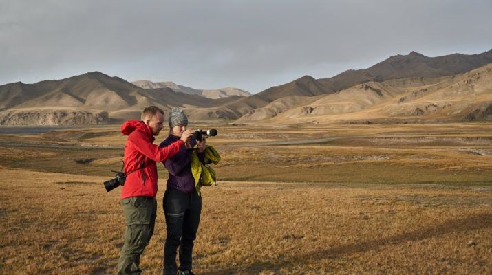 Two photographers taking pictures in a vast, open landscape with mountains in the background.